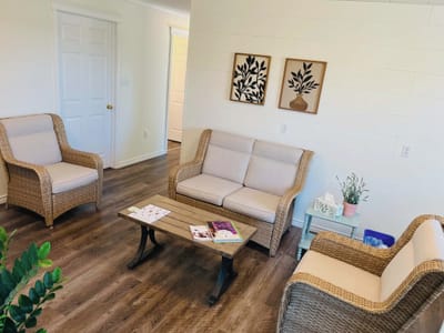 The waiting room: wicker chairs with cream cushions around a wooden coffee table, a small mint-green side table with a lavender plant, and two framed botanical prints on a white shiplap wall.