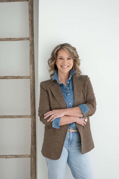 Lisa leaning against a white wall beside a rustic wooden ladder, arms crossed, smiling, in a brown tweed blazer and denim shirt.