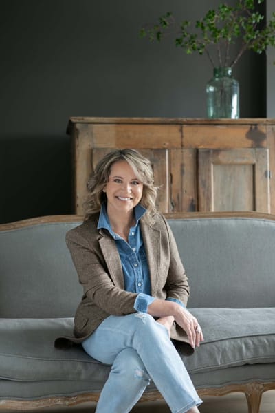 Lisa Fowler smiling, seated on a grey linen settee in front of a weathered pine cabinet.